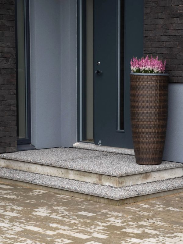 Pesubetoonplaatidega kaetud kaheastmeline välistrepp modernse maja sissepääsu juures, kõrge pruun lillepott roosade kanarbikega trepi serval.